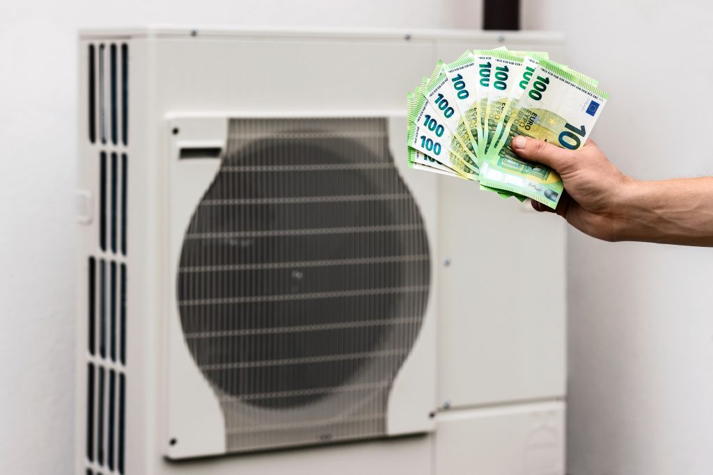 Wärmepumpe Kosten: Hand mit Geldscheinen vor Luftwärmepumpe.