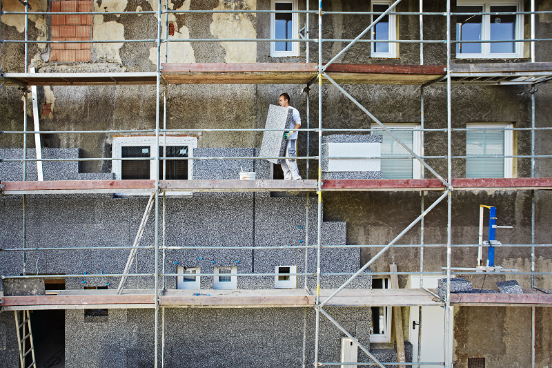 Handwerker geht auf einem Gerüst und trägt ein WDVS-Element für die Dämmung eines Altbaus.