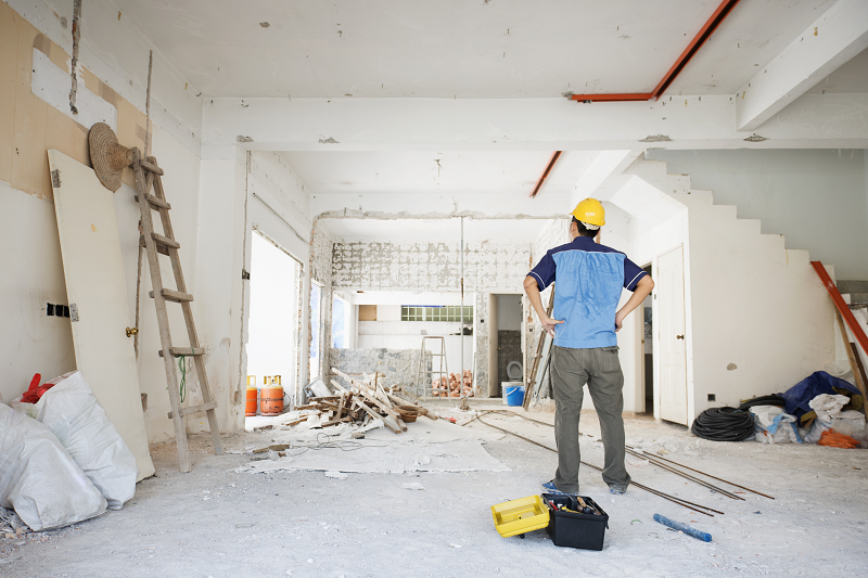 Handwerker steht mit eingestützten Händen und dem Rücken zur Kamera und betrachtet ein unsaniertes Zimmer.