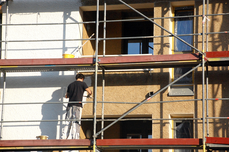 Mann steht auf einem Baugerüst und streicht eine ockerfarbene Fassade weiß.